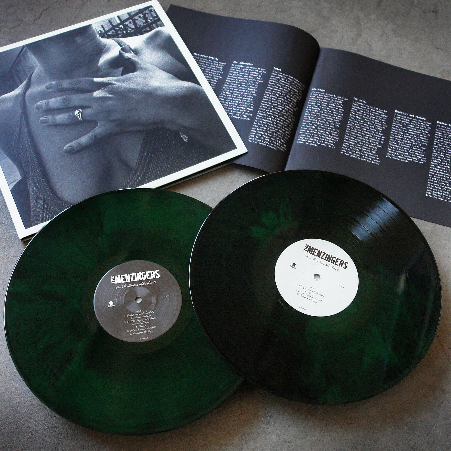 image of two neon green and black vinyls for the menzingers album on the impossible past on a concrete floor. the album cover is a black and white photo of a woman with her hand over her chest. the 8 page booklet is laid out to show the song titles and lyrics