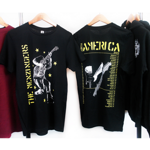 image of the front and back of a black tee shirt hanging on a white background. front of tee is on the left and has a full body print in yellow on the left side vertically says the menzingers with yellow stars all over and in white an image of a man playing a guitar. the back of the tee is on the right and has in yellow at the top 2022 america with the list of tour dates and locations below and a broken guitar in the center