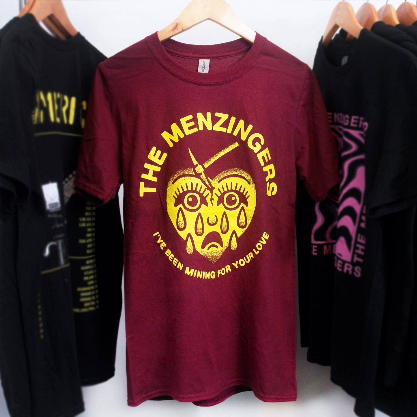 image of a maroon tee shirt hanging on a white background. the tee shirt has a full center chest print in yellow of a heart with a sad face and tears and a mining pick in the top. arhced around the heart says the menzingers. arhced around at the bottom of the heart says i've been mining for your love